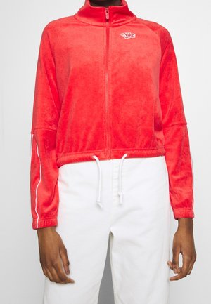 Persoon draagt een felrode Nike fleece-jas met rits en witte trekkoorden, gecombineerd met hoogwater witte broek tegen een effen achtergrond.