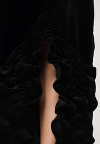 Svart fløyelsplagg med volanger, med en slisse opp til låret. Den myke teksturen og den glatte finishen fremhever designdetaljene.