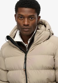 Beige pufferjack met een ritssluiting aan de voorkant, zwarte accenten, een capuchon met trekkoord en een logo-label. Gevoerde binnenvoering zichtbaar.