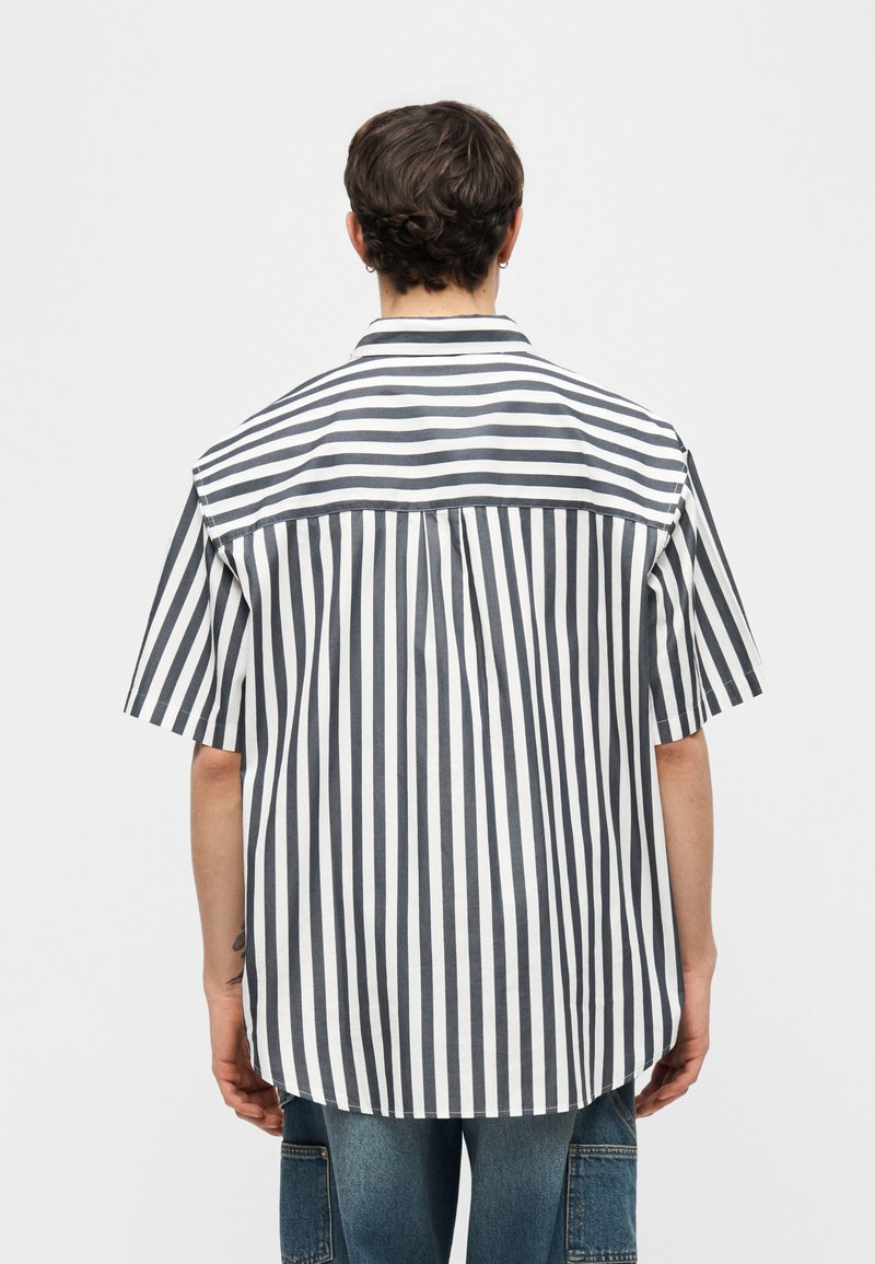 Persona che indossa una maglietta a maniche corte con strisce verticali e orizzontali nere e bianche, in piedi di spalle su uno sfondo semplice.