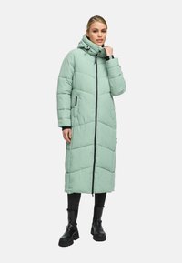 Woman wearing a long, mint green puffer coat with a hood, black pants, and black chunky boots, standing against a plain background.