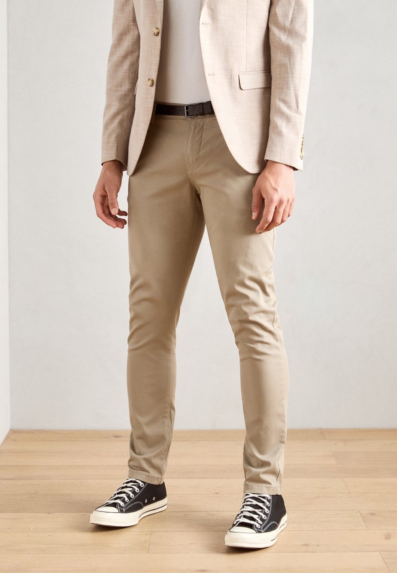 Homme portant un pantalon beige slim, un blazer beige clair et des baskets montantes noires, debout sur un sol en bois.