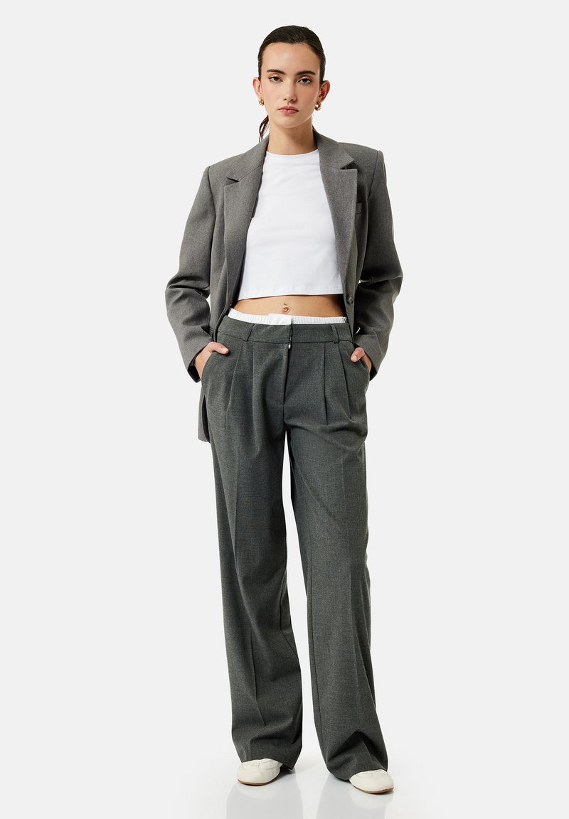 Femme debout, les mains dans les poches, portant une veste de costume grise, un pantalon large assorti, un top court blanc et des chaussures blanches sur fond uni.