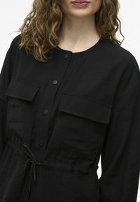 Čierna, ľahká košeľa s okrúhlym výstrihom, dvoma ná prsníkmi, predným gombíkovým zapínaním a detailom s nastaviteľným pásom. Hladká textúra.