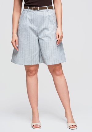 Kvinde iført lysegrå, stribede shorts med vide ben, brun bælte og beige åbne hæle mod en ensfarvet baggrund.