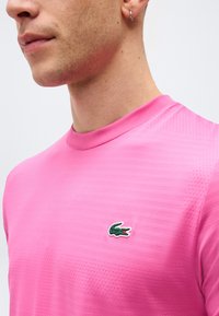 Άνδρας που φοράει φωτεινό ροζ πουκάμισο με υφή και μικρό πράσινο λογότυπο κροκοδείλου στο στήθος, με ασημένιο κρίκο στο αριστερό αυτί.