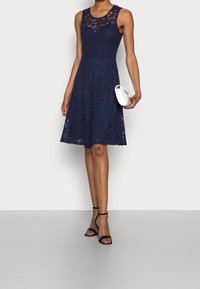 Marinblå spetsklänning med en figursydd liv och utsvängd kjol, med en genomsiktig spetsurringning. Matchad med en vit kuvertväska och svarta klacksandaler.