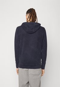 Navy blue hooded sweatshirt made from soft fabric, featuring a classic cut, ribbed cuffs, and a kangaroo pocket. Gray sweatpants visible below.
