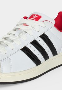 Λευκό αθλητικό παπούτσι Adidas με μαύρες ρίγες, κόκκινη φτέρνα και ετικέτα στη γλώσσα, λευκά κορδόνια και υφασμένο υπόλευκο πάτο.