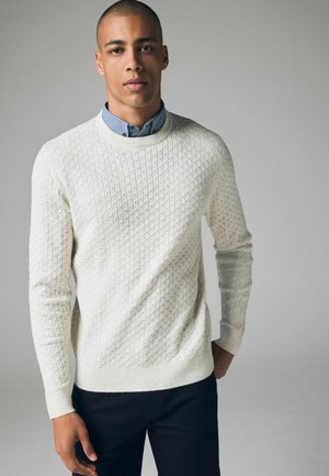 Suéter blanco de punto con un patrón texturizado, escote redondo y puños acanalados, combinado con una camisa de color azul claro con cuello debajo.