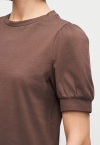 Chemise marron à manches courtes avec manches bouffantes et poignets ajustés portée par une personne à la peau claire sur un fond uni.