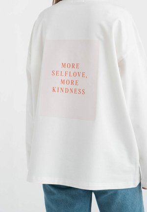 Personne portant un sweat-shirt blanc avec un texte orange dans le dos indiquant "MORE SELFLOVE, MORE KINDNESS" et un jean bleu.