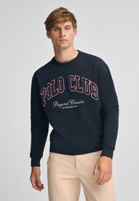 Mörkblå sweatshirt med "POLO CLUB"-logotyp i vinröd ovanför texten "Beyond Classics". Har rund halsringning och ribbade ärmslut och nederkant.