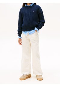 Pull en maille torsadée bleu marine, chemise à rayures bleu clair en dessous, pantalon large crème, baskets beiges avec semelles marron et détails en lacets.