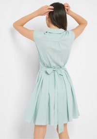 Light mint green linen dress with a fitted bodice, short sleeves, and a tied waist. Features pleated skirt and collar detail.