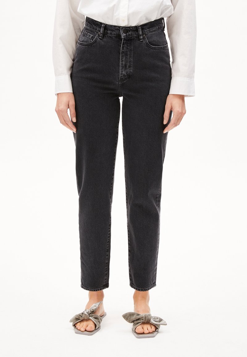 Personne portant un jean noir taille haute, une chemise blanche à manches longues et des sandales gris avec un nœud papillon, debout devant un fond blanc.