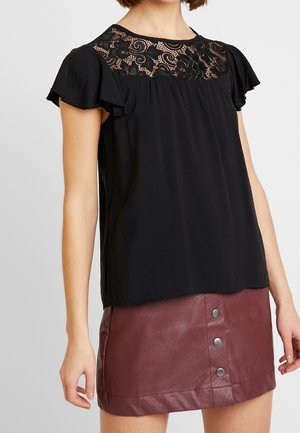 Vrouw draagt een zwarte kanten inzet blouse met wijde mouwen en een bordeauxrode lederen minirok met knopen aan de voorkant.