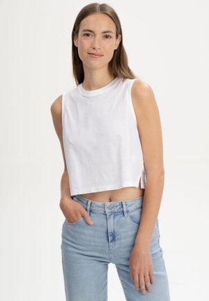 Frau mit langen braunen Haaren, die ein weißes ärmelloses, bauchfreies Top und hellblaue, hoch taillierte Jeans trägt, lächelnd mit einer Hand in der Tasche.