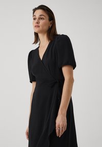 Black wrap dress with short puff sleeves, textured fabric, deep V-neck, and a tie waist for adjustable fit. Hem drapes elegantly.