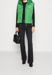 Zielona pikowana kamizelka z kapturem, zapinana na zamek błyskawiczny, z bocznymi kieszeniami, zestawiona z czarnymi rozkloszowanymi jeansami i czarną torebką.