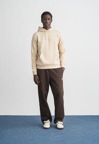 Bézs pamut kapucnis pulóver húzózsinórokkal és elülső zsebbel, sötétbarna melegítőnadrággal párosítva. Fehér logó a nadrágon. A modell kék padlón áll.