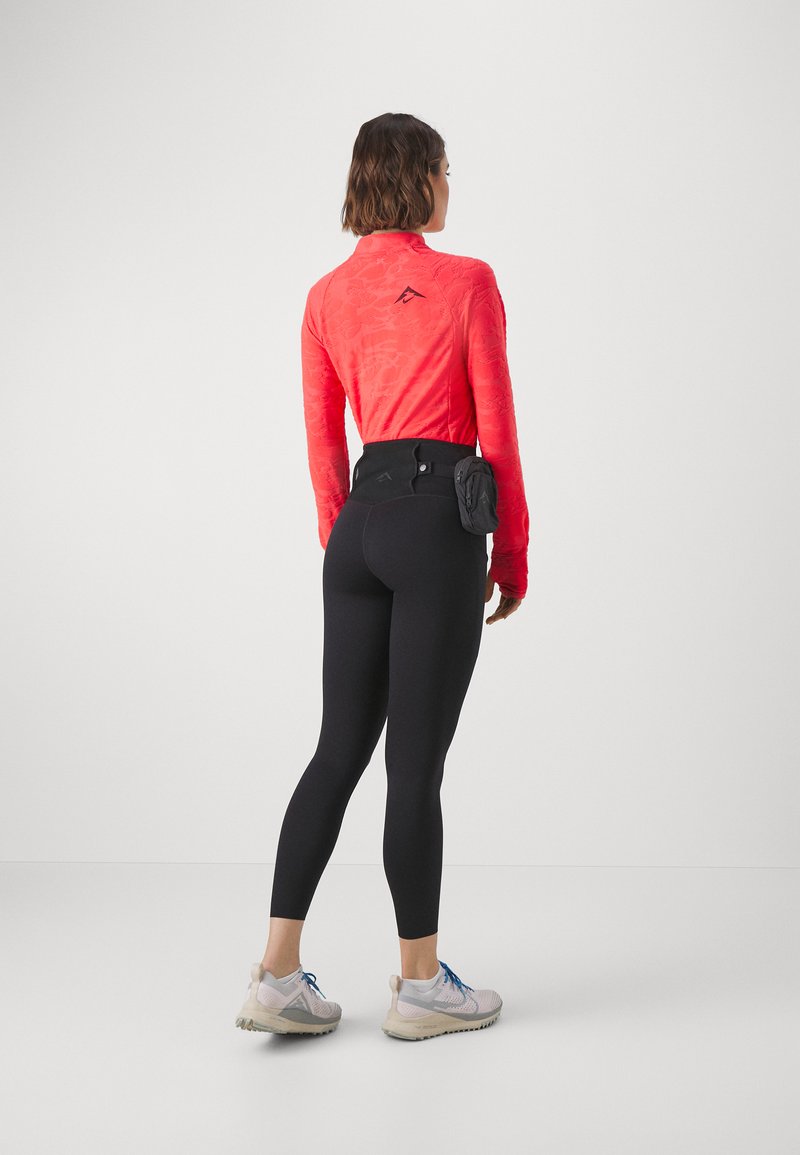 Top de sport à manches longues rouge avec une texture subtile, associé à des leggings noirs. Poche pour accessoires attachée à la taille. Baskets légères.