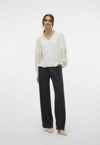 Witte blouse met getextureerde lange mouwen en kantaccenten, gecombineerd met hoge zwarte wijde broek en beige puntige schoenen.
