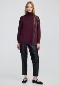 Kobieta stoi w burgundowym swetrze z golfem z skórzanymi paskami oraz czarnych skórzanych spodniach 3/4, w czarnych balerinach na jednolitym tle.