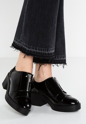 Czarne lakierowane buty na masywnym obcasie, z ozdobnym srebrnym zamkiem błyskawicznym i zaokrąglonym noskiem. Noszone z postrzępionymi czarnymi jeansami.