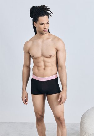 Άνδρας χωρίς πουκάμισο με dreadlocks, φοράει μαύρα εσώρουχα Lacoste με ροζ ζώνη, στέκεται σε ανοιχτό γκρι πάτωμα μπροστά σε απλή λευκή τοίχο.