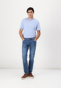 T-shirt bleu clair avec un petit logo en texte blanc, associée à un jean en denim bleu moyen et des baskets marron. Tenue simple et décontractée.