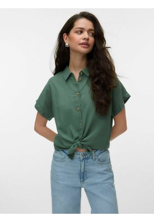 Woman with long dark hair wears green short-sleeve buttoned shirt tied at waist and light blue jeans, standing with hands behind back.