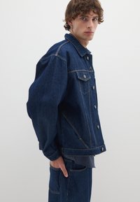 Giubbotto in denim di colore blu scuro con vestibilità rilassata, dotato di bottoni argentati, due tasche sul petto e dettagli di cucitura in vista.