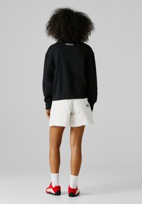 Oseba z kodrastimi lasmi, oblečena v črno trenirko, bele obrabljene kratke hlače, bele nogavice in rdeče superge, stoji obrnjena stran na sivi podlagi.