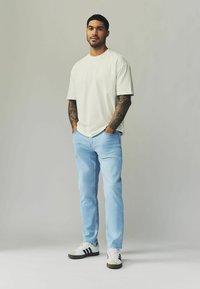 T-shirt a girocollo color beige chiaro, jeans dritti blu chiaro e sneakers bianche con strisce nere. Braccia tatuate visibili.