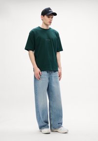 Mørkegrønn bomulls t-skjorte med logo foran, kombinert med lyseblå videnskjerte denimjeans og hvite sneakers. Modell står oppreist.