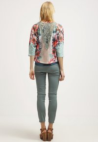 Bluse mit Blumenmuster und Dreiviertelärmeln, Häkelrücken, kombiniert mit engen grünen Hosen und klobigen braunen Absätzen.