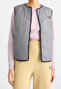 Chaleco aislante gris con cierres a presión, ribete negro y detalle de logo. Lleva puesto sobre una camisa rosa clara y pantalones beige texturizados.
