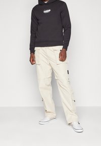 Felpa nera con cappuccio e logo bianco, abbinata a pantaloni cargo beige con tasche laterali, indossata con scarpe slip-on a scacchi.