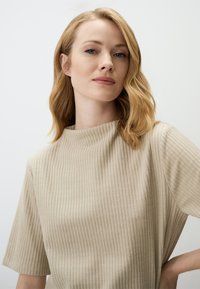 Top beige a costine con maniche corte e scollo alto. Il tessuto ha una consistenza morbida e delicate linee verticali, con una vestibilità rilassata.