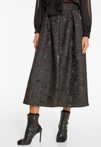 Zwarte gemusterde midi-jurk gecombineerd met een transparante blouse met lange mouwen en zwarte leren enkellaarsjes met hoge hakken en open tenen.