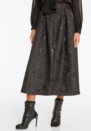 Jupe midi noire à motifs, associée à un blouse transparente à manches longues et des bottines en cuir noires à talons hauts avec bouts ouverts.