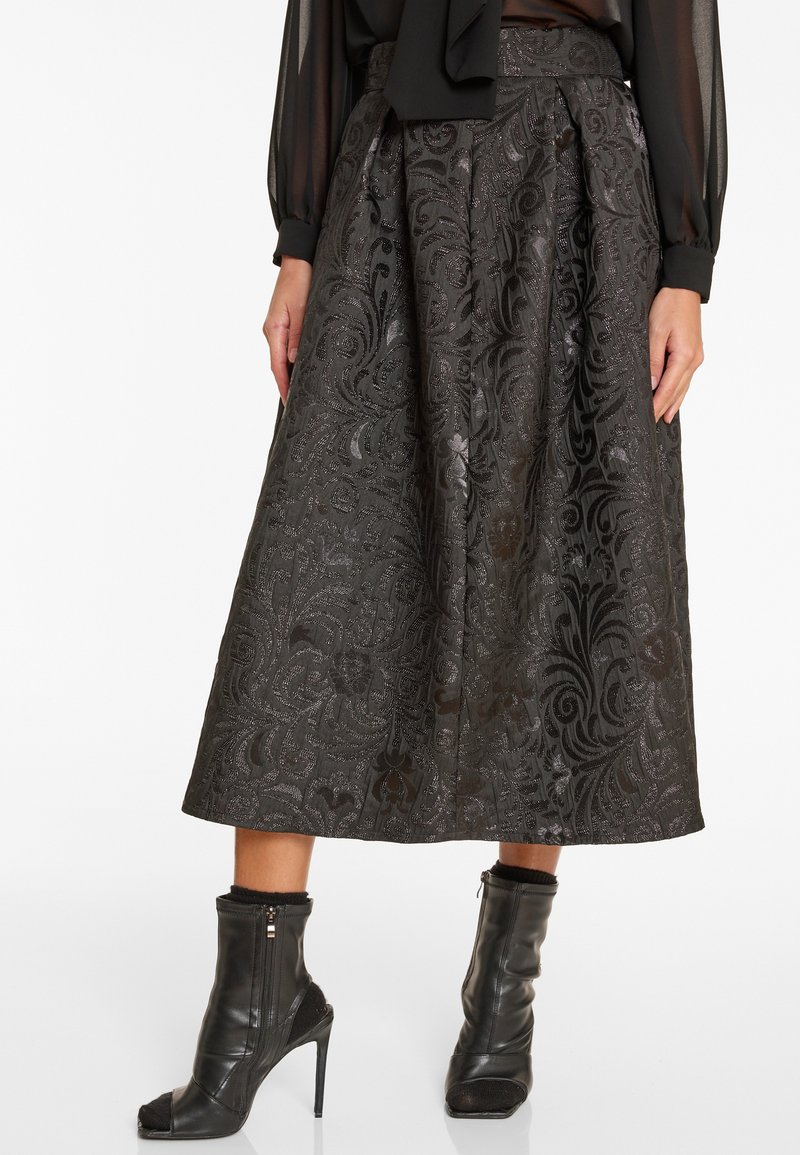 Zwarte gemusterde midi-jurk gecombineerd met een transparante blouse met lange mouwen en zwarte leren enkellaarsjes met hoge hakken en open tenen.