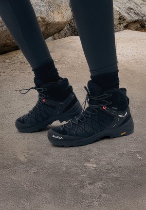 Czarne buty do wędrówek z wysokim kołnierzem, teksturowaną powierzchnią, sznurowaniem i podeszwą Vibram. Na bokach znajdują się czerwone i białe akcenty.