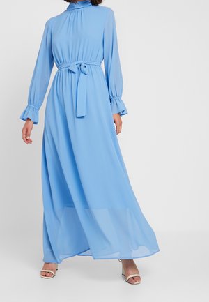 Kobieta ubrana w długą, lekko błękitną, zwiewną sukienkę z wysokim kołnierzem, długimi rękawami z falbanami przy mankietach i paskiem w talii, w połączeniu z białymi sandałami na otwartym palcu.