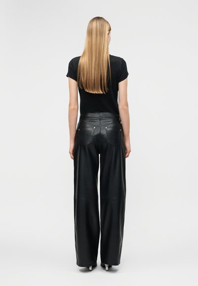 Γυναίκα με μακριά ξανθά μαλλιά στέκεται με την πλάτη, φοράει μαύρο τοπ με κοντά μανίκια και μαύρο δερμάτινο παντελόνι με ψηλή μέση και φαρδιά μπατζάκια.