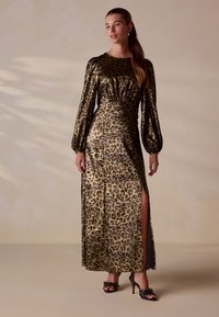 Maxikleid mit Leopardenmuster in metallischem Gold, langen Puffärmeln, vorderem Schlitz und tailliertem Schnitt. Kombiniert mit schwarzen Riemchen-High Heels.