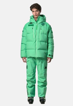 Giovane uomo in piedi che indossa una giacca imbottita verde menta con cappuccio, pantaloni da sci abbinati e scarponi da sci neri su sfondo semplice.