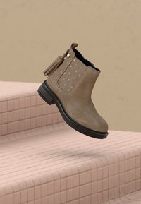 Botte de cheville en daim marron avec semelle noire, perforations en forme d'étoile sur le côté et détail de pampille, positionnée au-dessus d'escaliers carrelés.