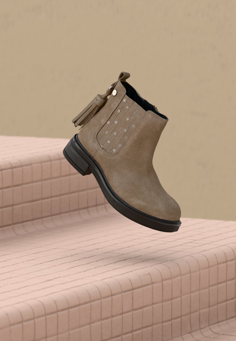 Botte de cheville en daim marron avec semelle noire, perforations en forme d'étoile sur le côté et détail de pampille, positionnée au-dessus d'escaliers carrelés.